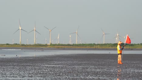 Right-side-composition,-a-coastal-guard-holding-a-red-flag,-standing-still-on-the-tidal-flats-with-wind-turbines-spinning-and-rotating-along-the-horizon-at-twilight,-Gaomei-wetland,-Taichung,-Taiwan