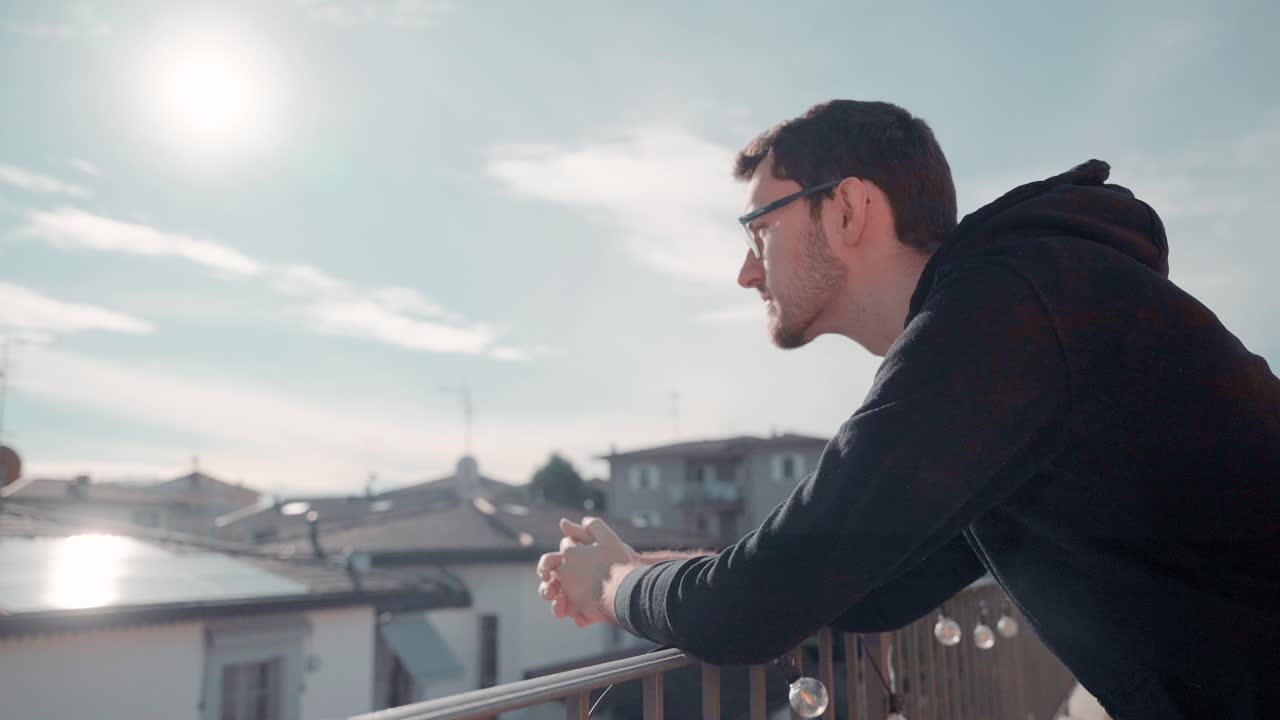 Jovem atraente caucasiano com barba e óculos na varanda, sob o sol, observando os arredores e desfrutando do bom tempo ensolarado