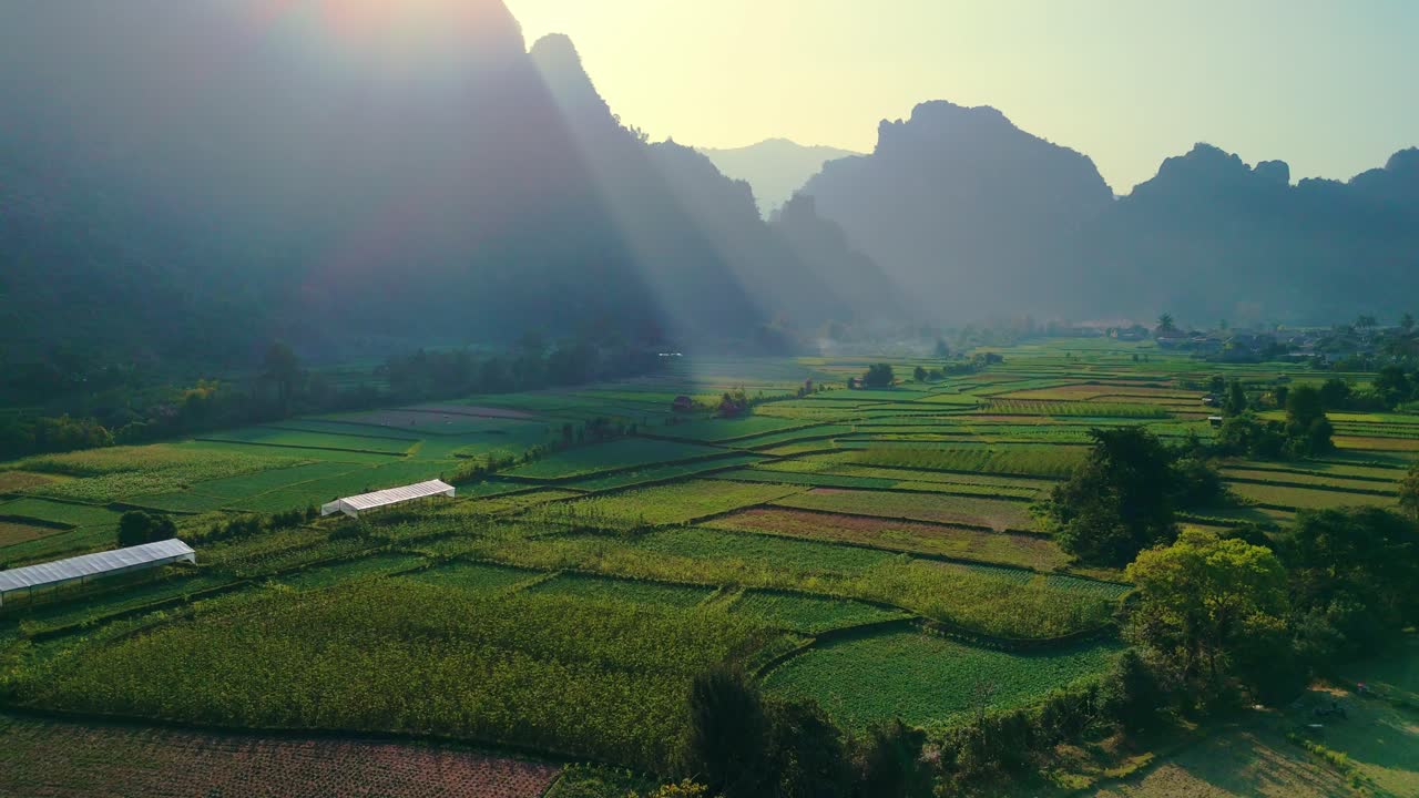 Mistica valle fertile e coltivazioni a mosaico sorvolano uno stagno riflettente, con i morbidi raggi del sole serale all'orizzonte montuoso