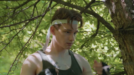 young man tying headband under low tree branches with elbows brushing thin twigs and sunlit leaves around as he prepares outdoor headwrap in dense green forest close up shot capturing focus