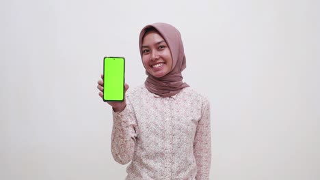 happy young asian muslim woman standing while presenting a blank green screen cell phone display