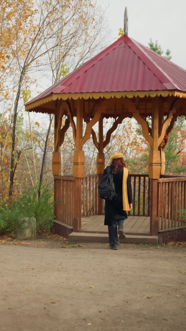 Back view of lady wearing black coat, yellow scarf, and beret walking into a wooden gazebo in a peaceful park, wooden bench nearby adds to the tranquil autumn setting surrounded by golden trees