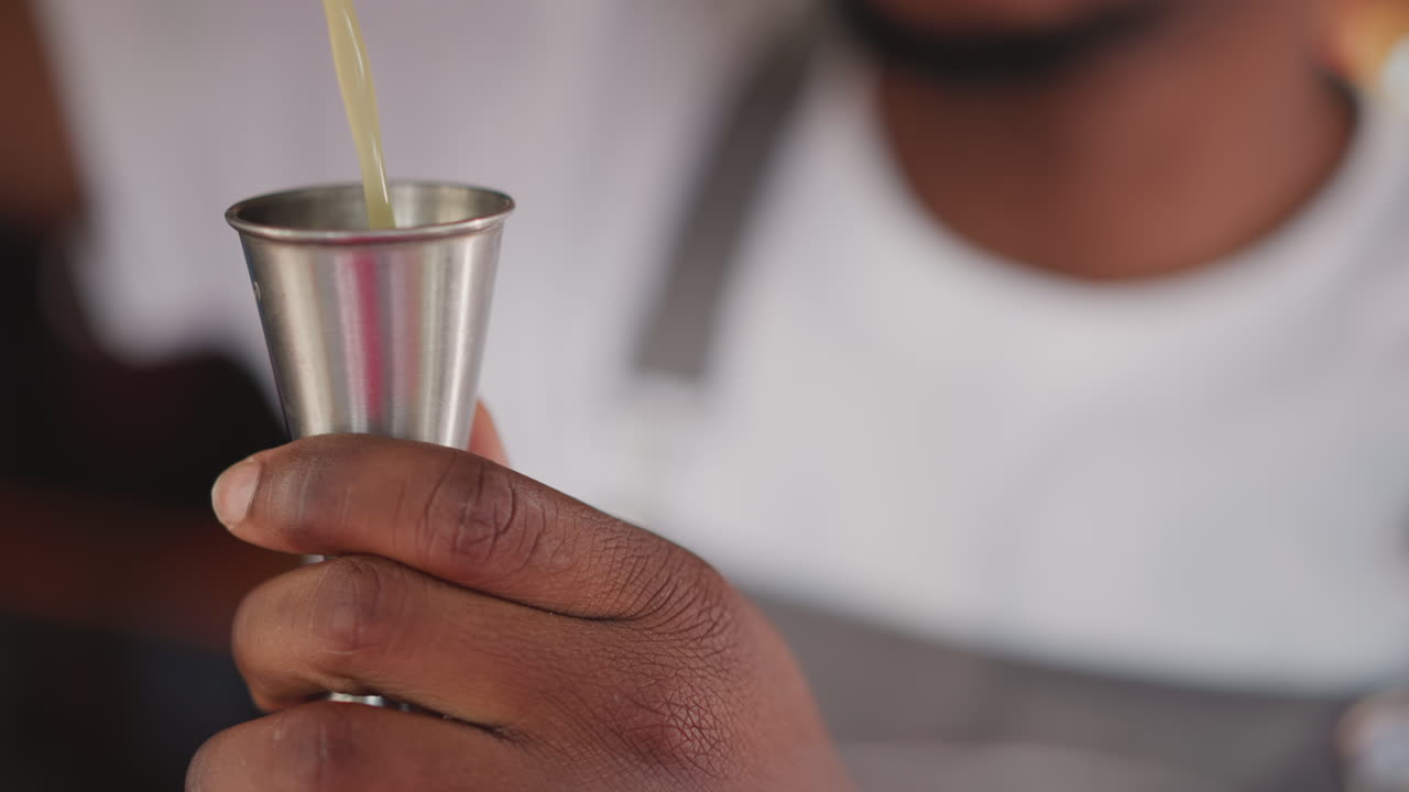 el camarero vierte jugo en un jigger inoxidable en un bar de primer plano. el camarero afroamericano mide los ingredientes líquidos para el cóctel. el hombre negro prepara una bebida exótica