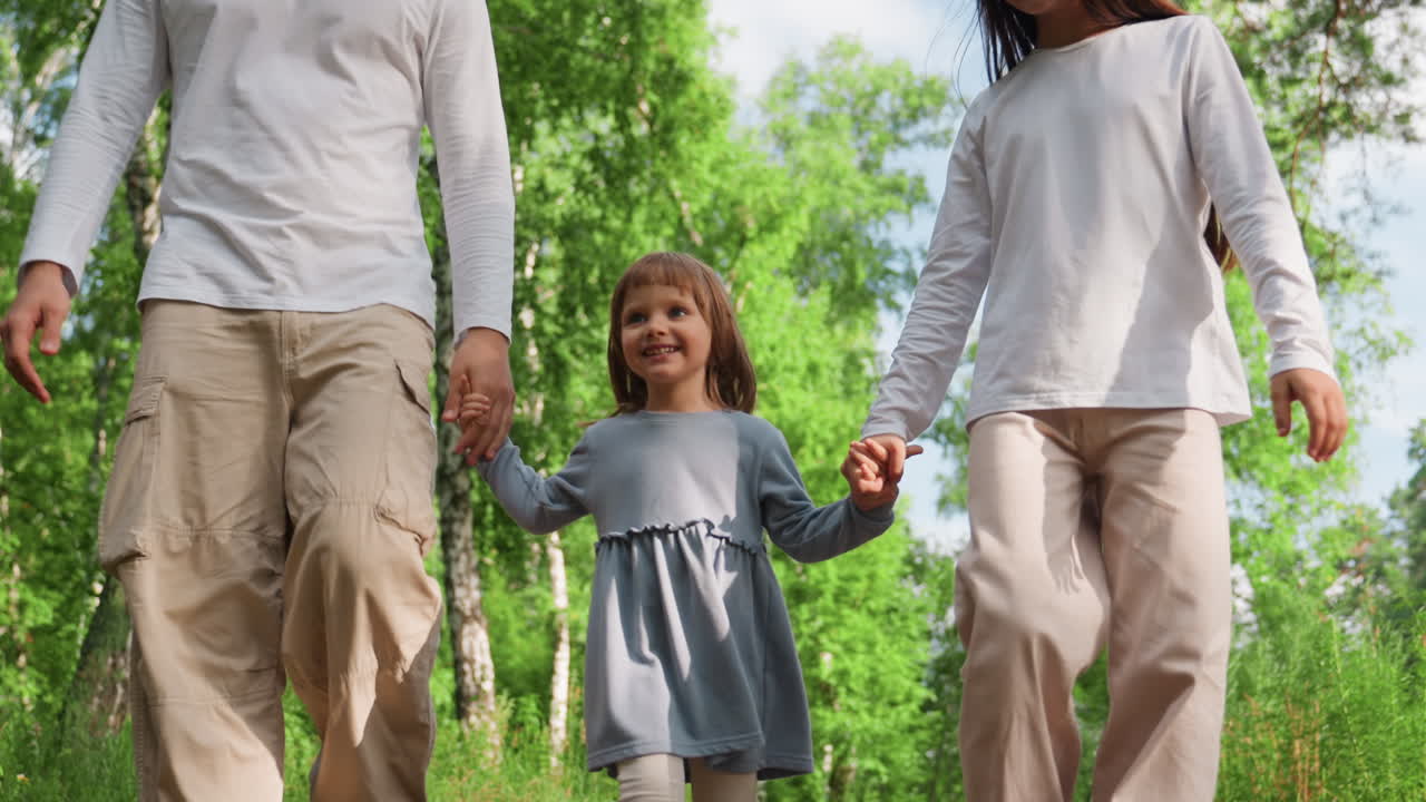 Close up of cheerful little girl walking between older siblings holding hands along forest path under sunlight, smiling with joy while enjoying nature and family warmth during peaceful outdoor stroll