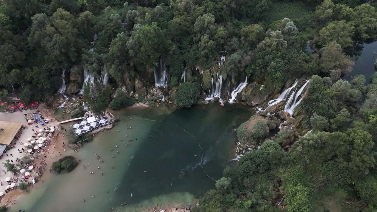Folle nuotano e si rilassano lungo la riva sabbiosa sotto le cascate a più livelli di Kravica, circondate da una fitta foresta verde e caffè lungo il fiume, viste da una prospettiva aerea con drone