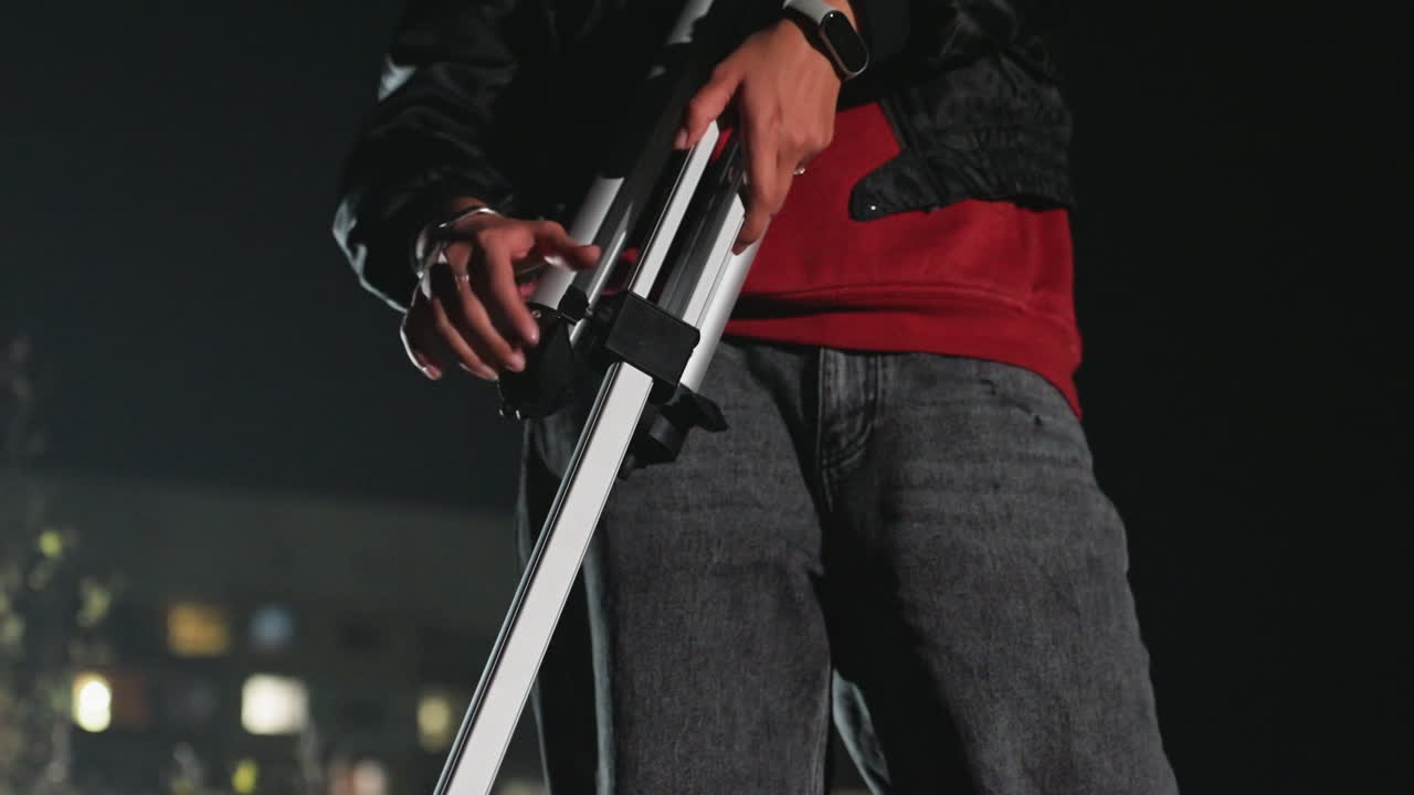 Astronomo sul tetto che regola il telescopio di notte, uomo con maglione rosso e giacca abbassa il telescopio bianco sul treppiede con luci della città sfocate sullo sfondo, primo piano a figura intera che mostra mani, orologio e jeans