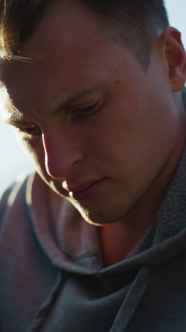 Close-up of young man outdoors wearing dark gray hooded sweatshirt, looking down with concentrated, pensive expression. Blurred green foliage in background