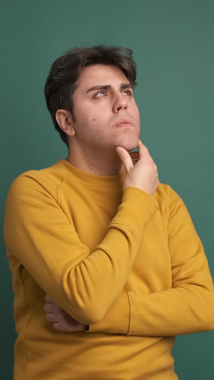 Thoughtful young man touching chin in green studio