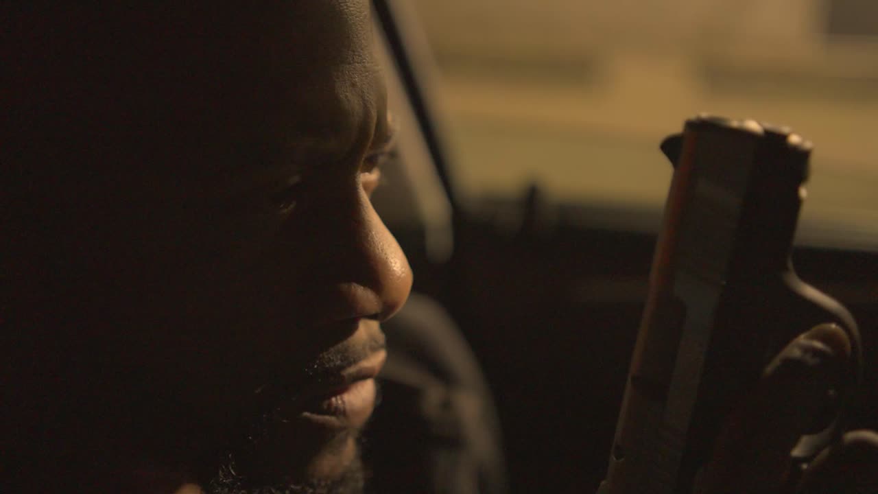 чернокожий мужчина, сидящий на сиденье водителя машины, рассматривает свой пистолет и ствол боевых патронов, прежде чем загрузить пули в свой пистолет для решения, меняющего жизнь