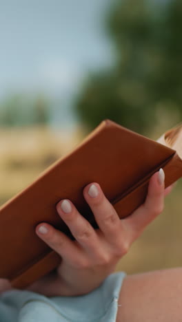 vista lateral de primer plano de una mujer leyendo un libro viejo al aire libre, volviendo páginas con manos delicadas, borrón suave en pantalones cortos azules y fondo rústico