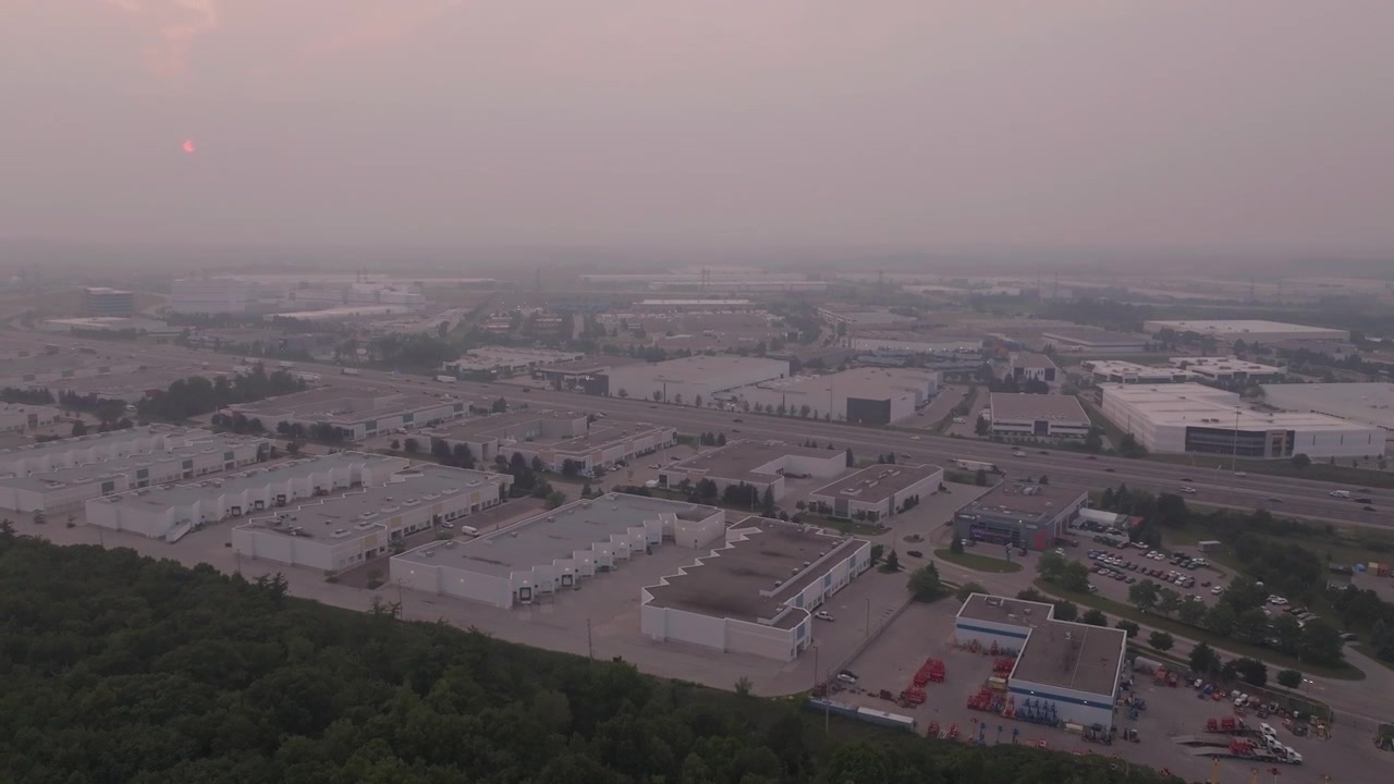 Una vista aérea de dron de ángulo alto revela zonas industriales cerca de la Autopista 401, oscurecidas por el denso humo de un incendio forestal. La atmósfera brumosa reduce la visibilidad sobre almacenes y estacionamientos vacíos.