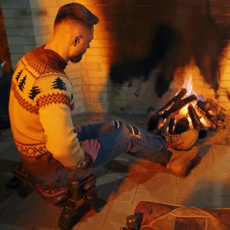 man is sitting near the fireplace. young male is warming hands near the fire inside. cozy evening by the fireplace at home. cold season concept.