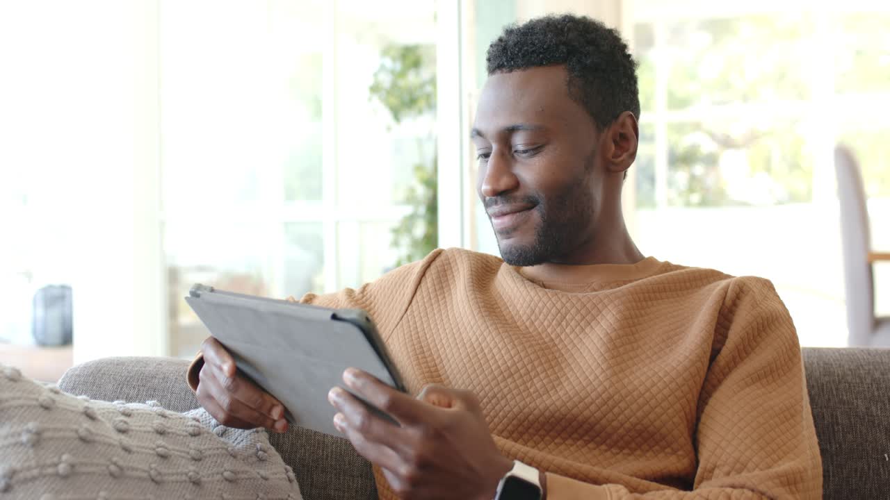 African American man scrolling tablet after notification popping up on living room couch, smiling