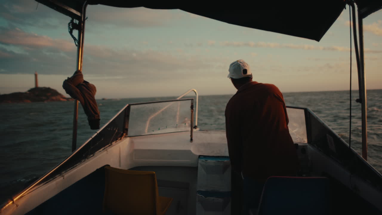 Man standing at front of small boat navigating across ocean toward distant coastline during sunset