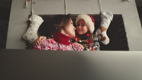 niña amorosa besando a su madre feliz mientras se sientan juntos en el sofá y usan una computadora portátil el día de navidad