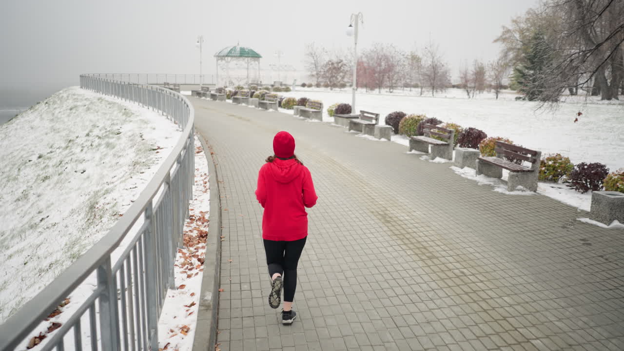 Back view of lady jogging in red beanie and jacket along snowy pathway near iron railing with hair fluttering as she moves, foggy atmosphere, decorative structure, light pole, and benches