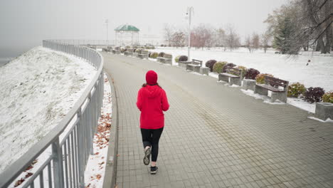 back view of lady jogging in red beanie and jacket along snowy pathway near iron railing with hair fluttering as she moves, foggy atmosphere, decorative structure, light pole, and benches