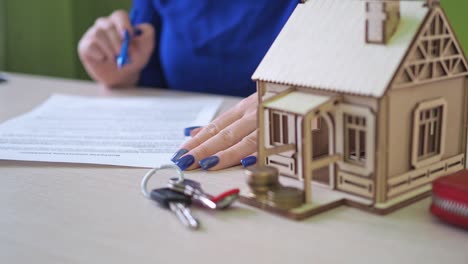 a woman signs a mortgage agreement for a house. there is a wooden house on the table, the keys to the property. familiarization with the purchase agreement