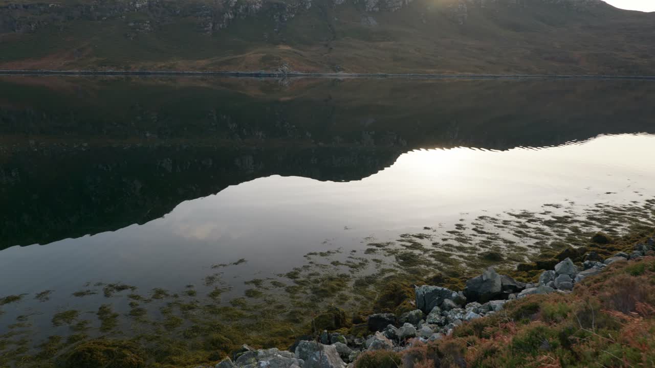 una toma panorámica lenta de agua tranquila en un lago marino en escocia mientras el sol se pone detrás de las montañas