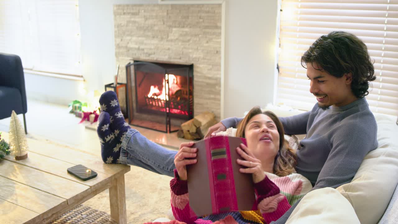 Diverse couple turning page in red book, reading aloud on sofa by fireplace for cozy moment