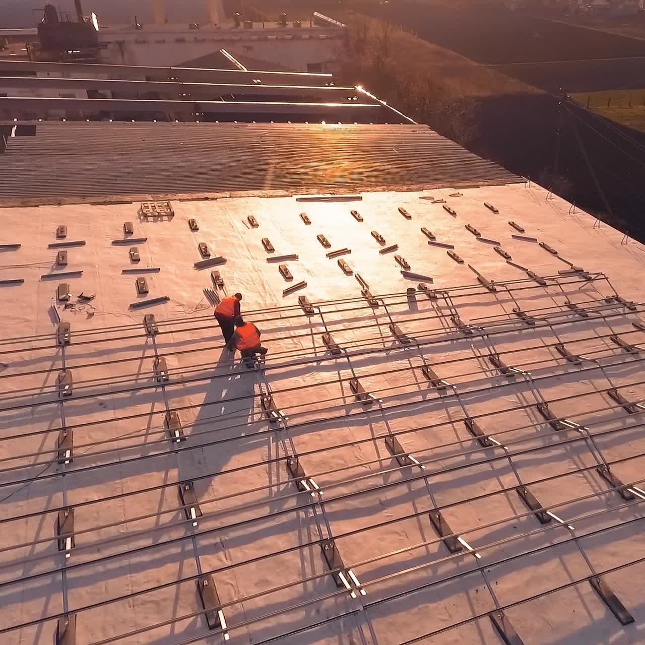 Installation solar farm on a roof at sunset. Workers in orange uniform installing metal basis on the roof for solar panels. Aerial view.