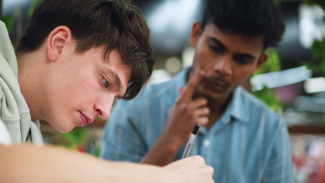 Indian boy with fluffy hair seated at wooden table scratching cheek while attentively watching friend sketch focused detailed drawing on paper under soft light in collaborative art session