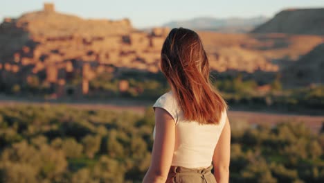 Close-view-of-young-caucasian-woman-walking-and-looking-around-through-desert-landscape-at-sunrise-in-Ait-Ben-Haddou,-Morocco