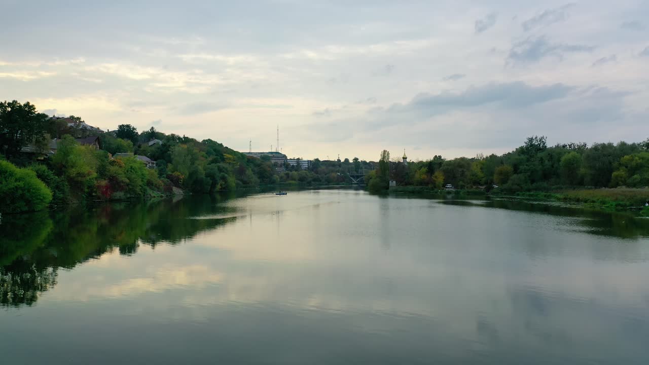 Calm water in the evening. River among green trees from both sides. Panoramic view of a river in the town. Motion camera back. Aerial view.