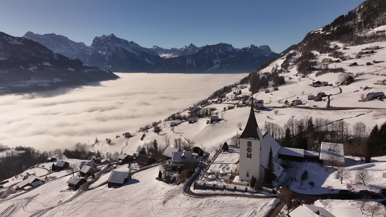 Stunning aerial view of snow-covered Swiss village, church and cloud covered Walensee, Amden, Switzerland with breathtaking alps in background during winter.