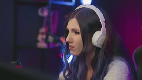 curious female gamer wearing white headphones seated in colorful studio reacts with displeasure after losing game, leaning forward with focused and serious expression under bright ambient lighting