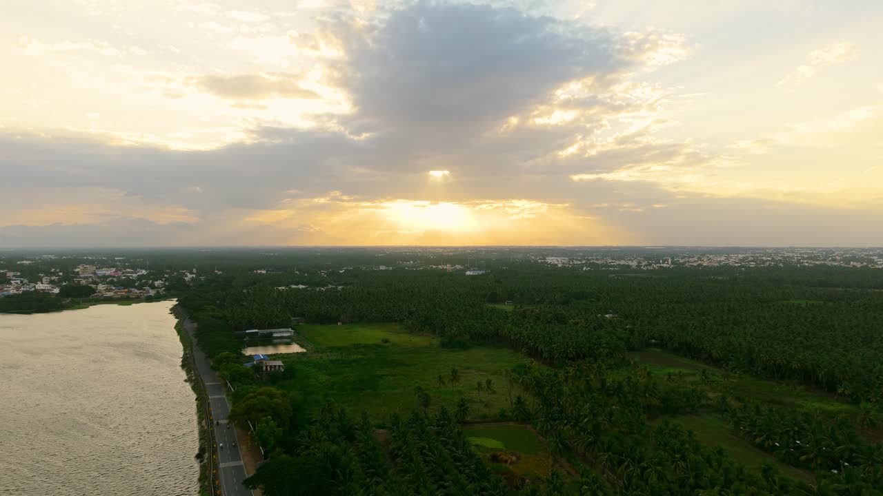 Cinematic aerial view of sunset spreads golden rays over green fields and a calm lake, with dramatic clouds coloring the sky and creating a breathtaking evening view of nature’s beauty