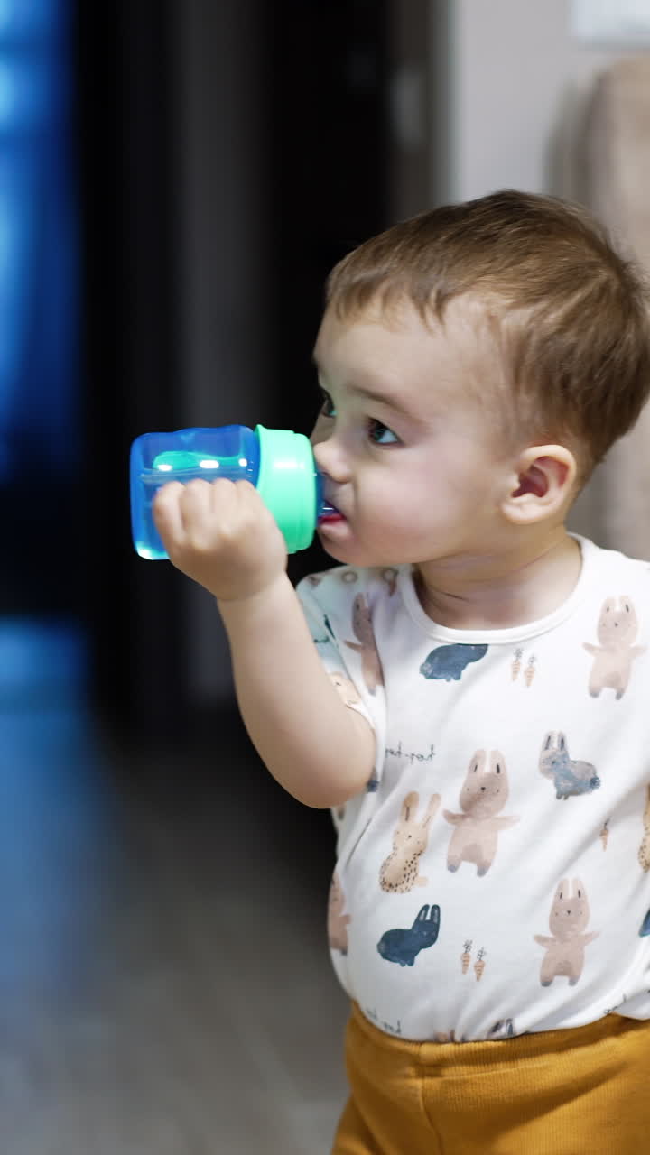Little child stands in the room drinking water from a bottle. Lovely boy drops the bottle and approaches the camera. Vertical video