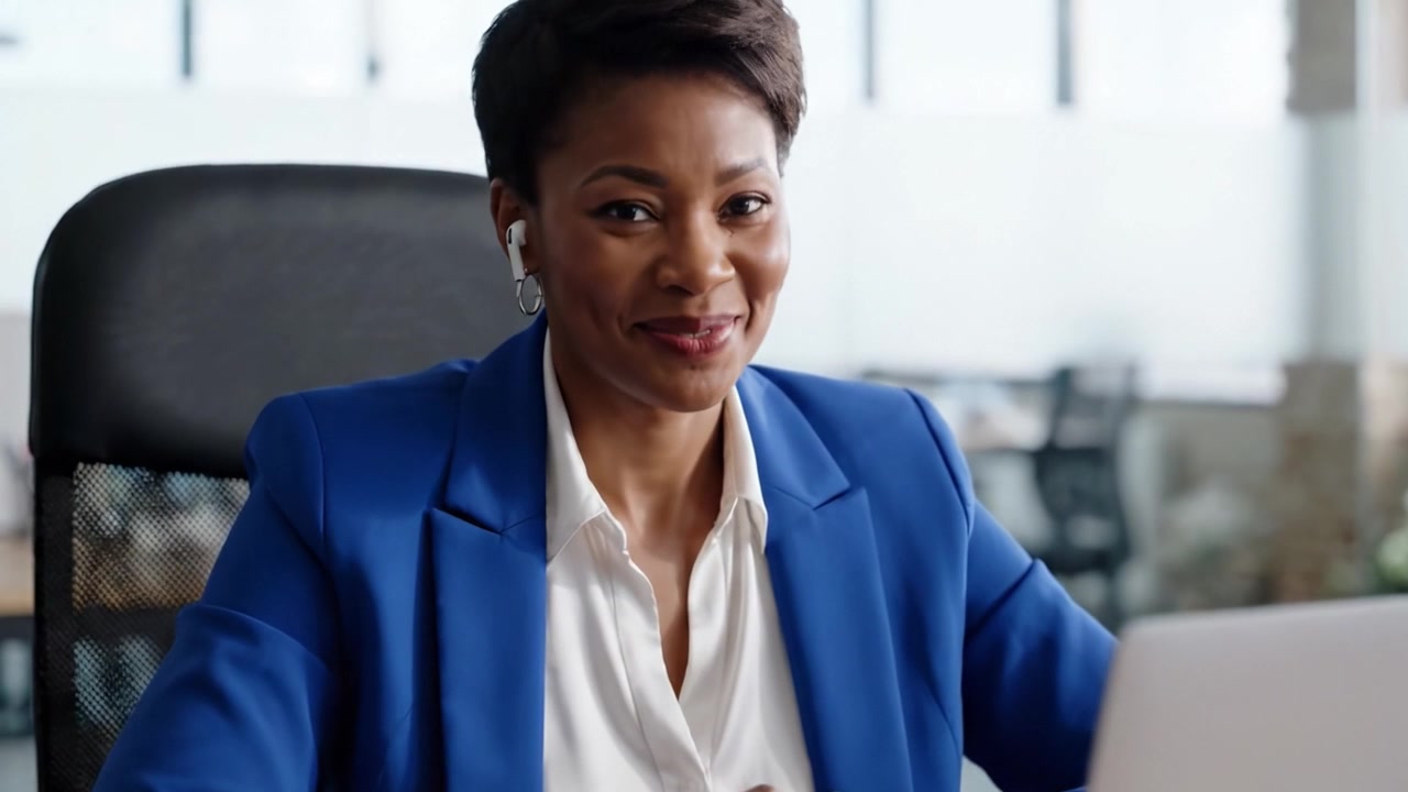 A confident woman in a blue blazer engages with her laptop, showcasing a positive attitude and focus in a modern office environment during a video call or online meeting.
