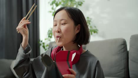 young asian woman in headphones eats noodles passionately with wood sticks at home