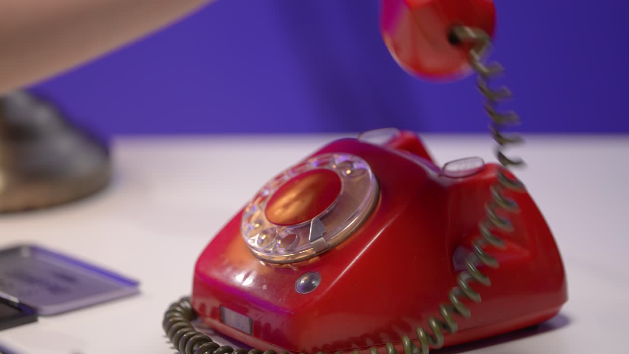 A delicate hand rests on a red rotary telephone in a vintage-inspired office setting. The nostalgic tone, bold colors, and retro props evoke timeless communication and thoughtful stillness