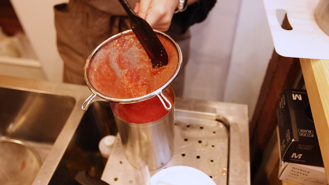 Un cuoco con un grembiule marrone filtra della purea di frutti rossi in un contenitore di acciaio inossidabile.