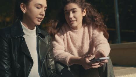 young female friends talking and using a smartphone outdoors