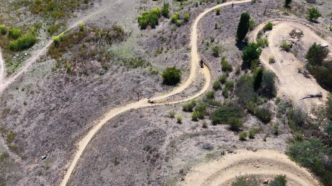 Plano aéreo em ângulo alto com drone acompanha um ciclista pedalando por uma trilha de terra sinuosa em região de mata
