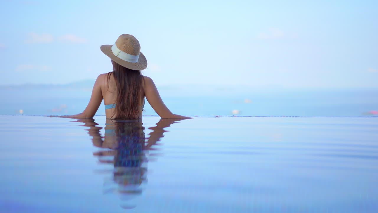Eine junge Frau mit dem Rücken zur Kamera verweilt am Rand eines Infinity-Pools und blickt auf das Meer.