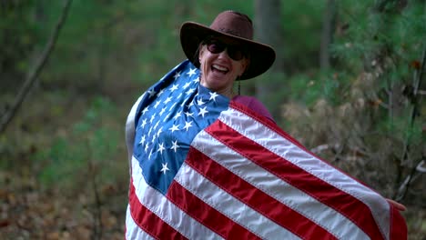 woman in cowboy hat and sunglasses holding out an american flag and holding it up to her body and laughing