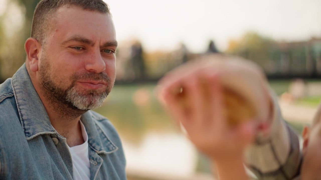 Primer plano de hombre con chaqueta vaquera y niño al aire libre cerca del agua y un puente. El niño ofrece comida al adulto, ambos interactuando juntos. La luz natural suave ilumina la escena con un fondo difuminado.
