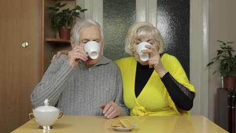 Happy-old-grandparents-couple-sit-on-table-at-home-enjoy-drinking-tea-together