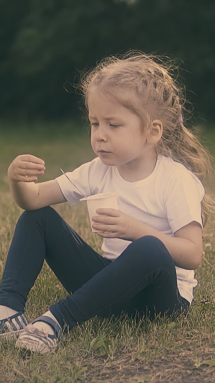 little pretty lady with long fair hair eats tasty yogurt sitting on grass in garden against green trees slow motion