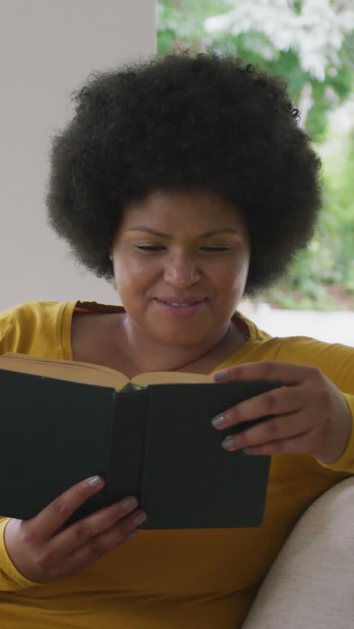 video de una feliz mujer afroamericana de tamaño grande acostada en el sofá y leyendo un libro