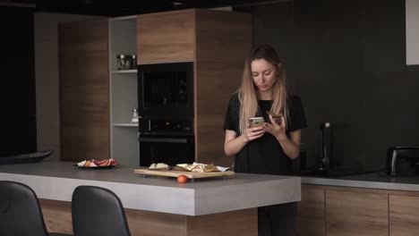 concentrated woman eating bread with butter for breakfast, checking her smartphone in kitchen