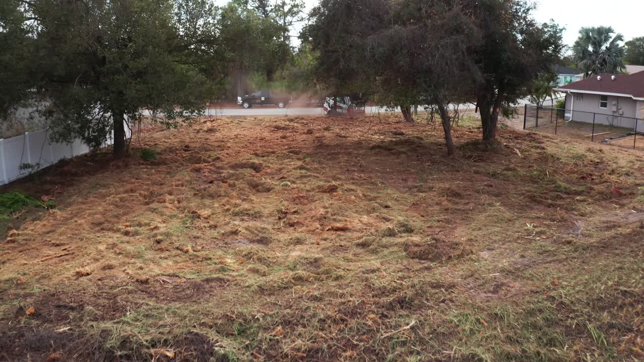 Un terreno recientemente despejado con árboles dispersos, bordeado por una calle tranquila. Pasan vehículos, indicando actividad residencial cercana. Útil para contenido de desarrollo urbano.
