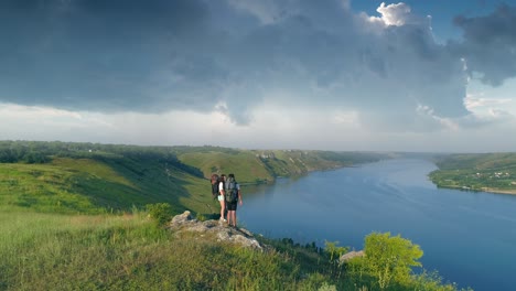 the two travelers with backpacks standing on the mountain top above scenic river