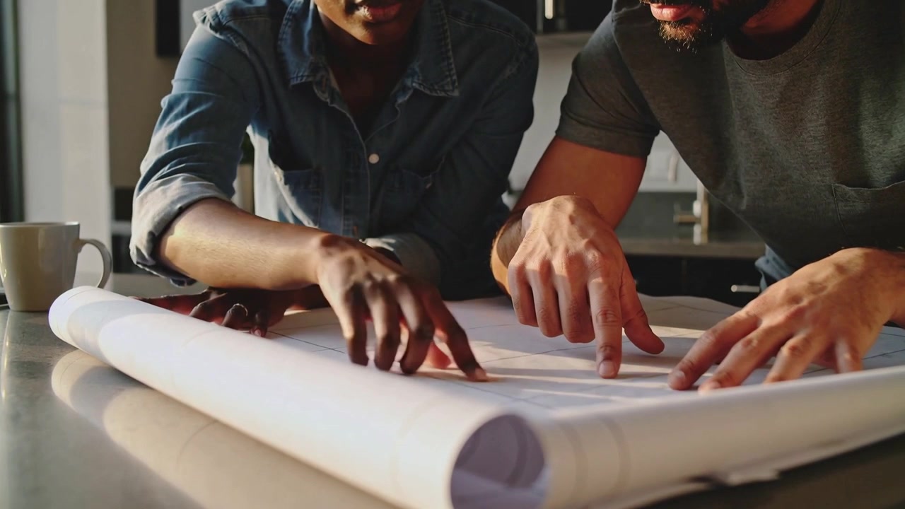 Unrolling couple leaning in kitchen, denim woman, bearded man tracing plans, finalizing plan, mug