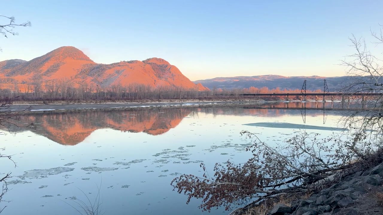 カムループスのマウントポールにて、澄み切った空の下、周囲の山々と橋を映し出す穏やかな川の静止画。自然の美しさと静けさを表現。
