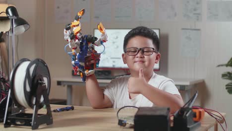 smiling asian cute boy with a cyborg hand showing thumbs up gesture to the camera while working on a laptop at home